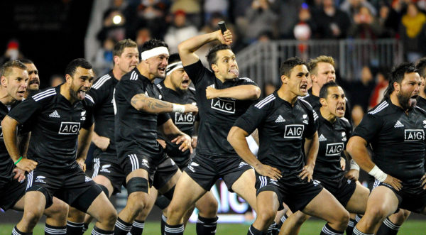 Giocatori della squadra di rugby degli All Blacks mentre mimano la celebre Haka, danza rituale maori: spalancano gli occhi, digrignano i denti, mostrano la lingua, si battono violentemente il petto e gli avambracci, esprimendo così potenza e coraggio, elementi propri dello spirito guerriero dei Maori.